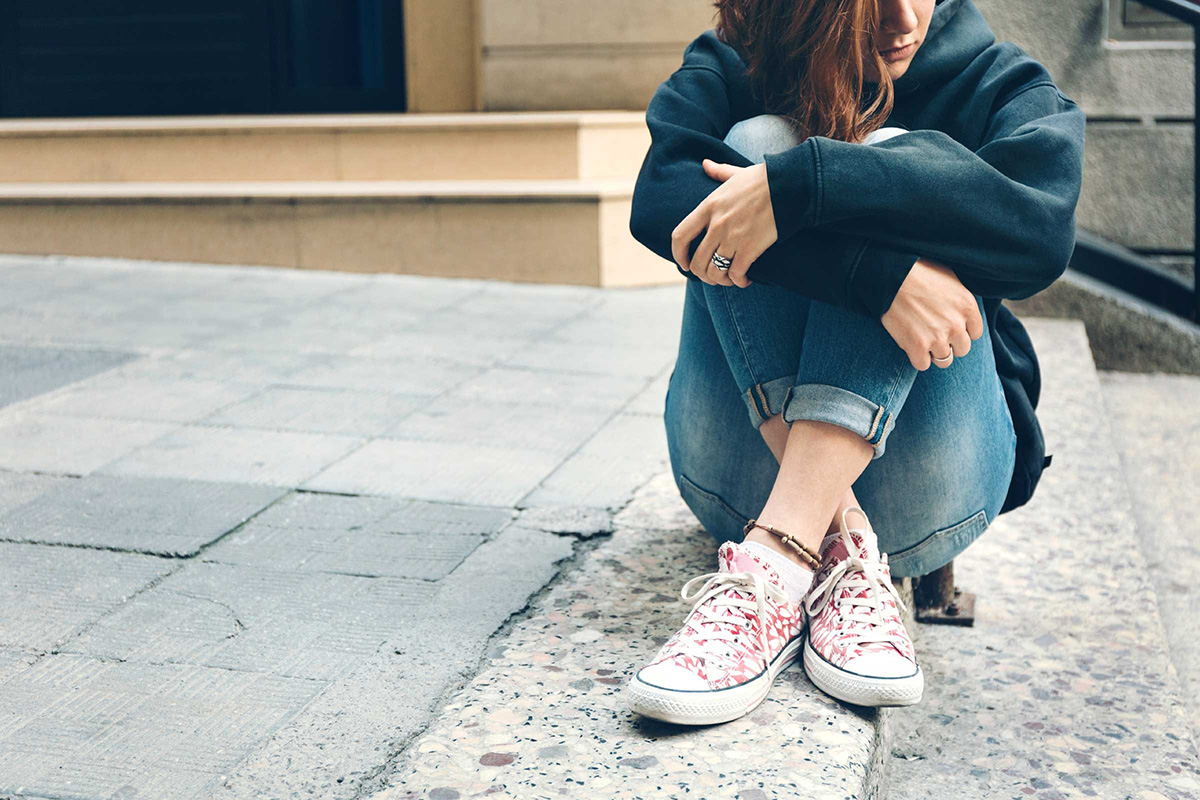 A teen sits on the ground with their knees pulled to their chest and looks off into the distance.