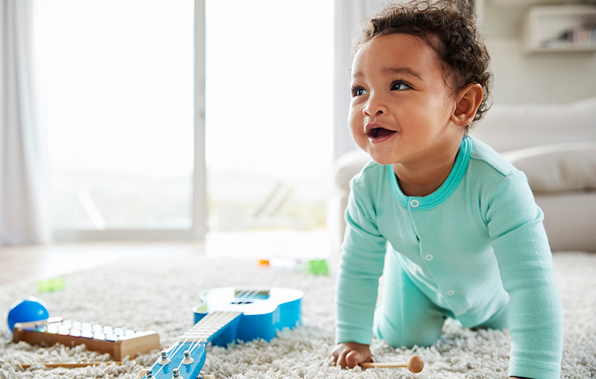 A baby in a mint green onesie smiles and crawls on a carpet scattered with toys.