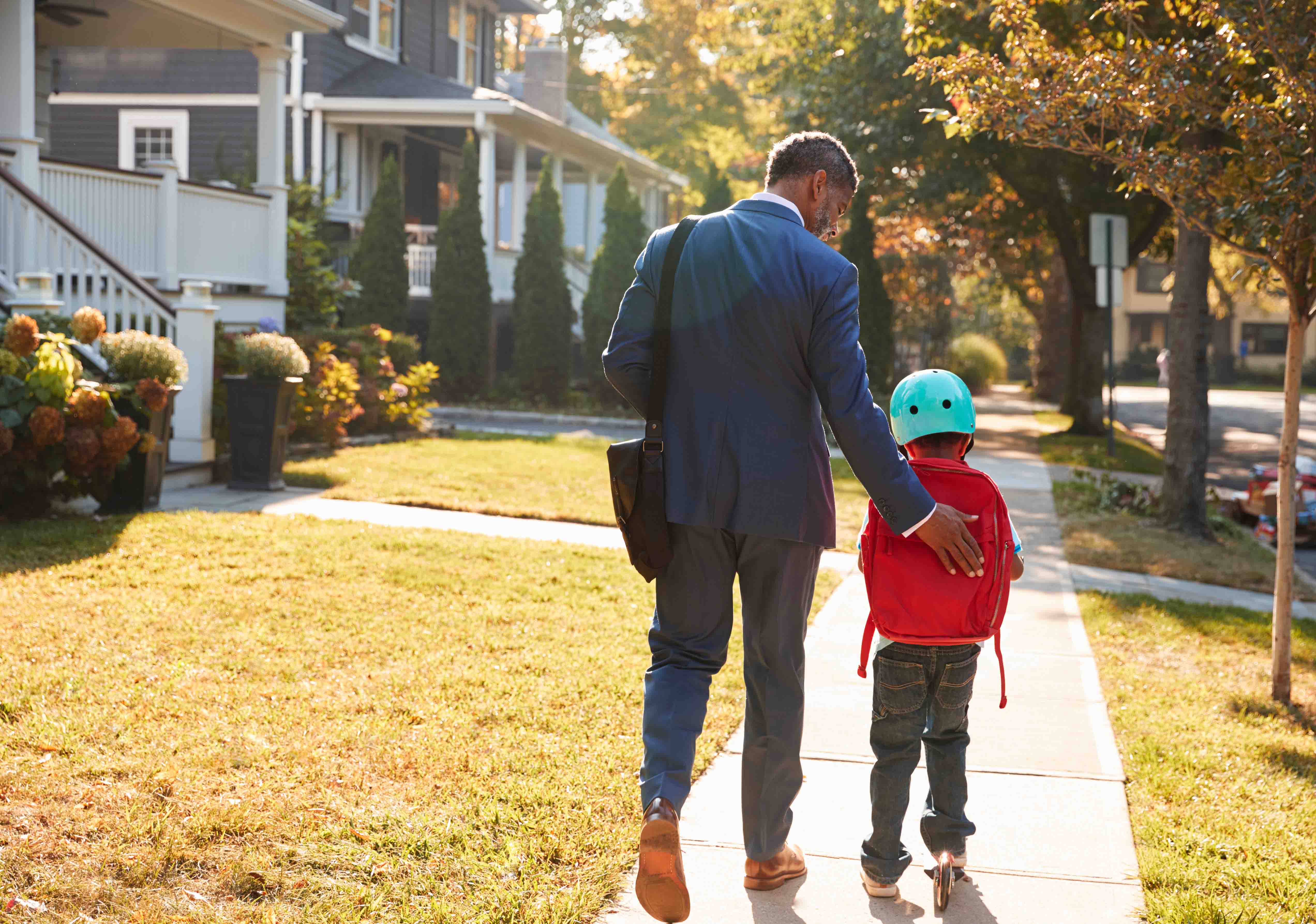 A man walks alongside a child on a sidewalk, sharing a moment together in a pleasant outdoor setting.